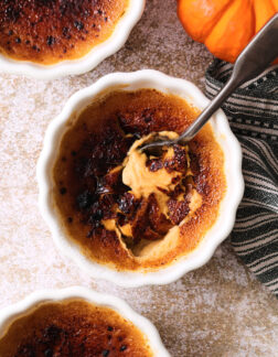 small white ramekin of pumpkin creme brulee, the burnt sugar crust has been cracked by a spoon and the pumpkin custard is just visible underneath.