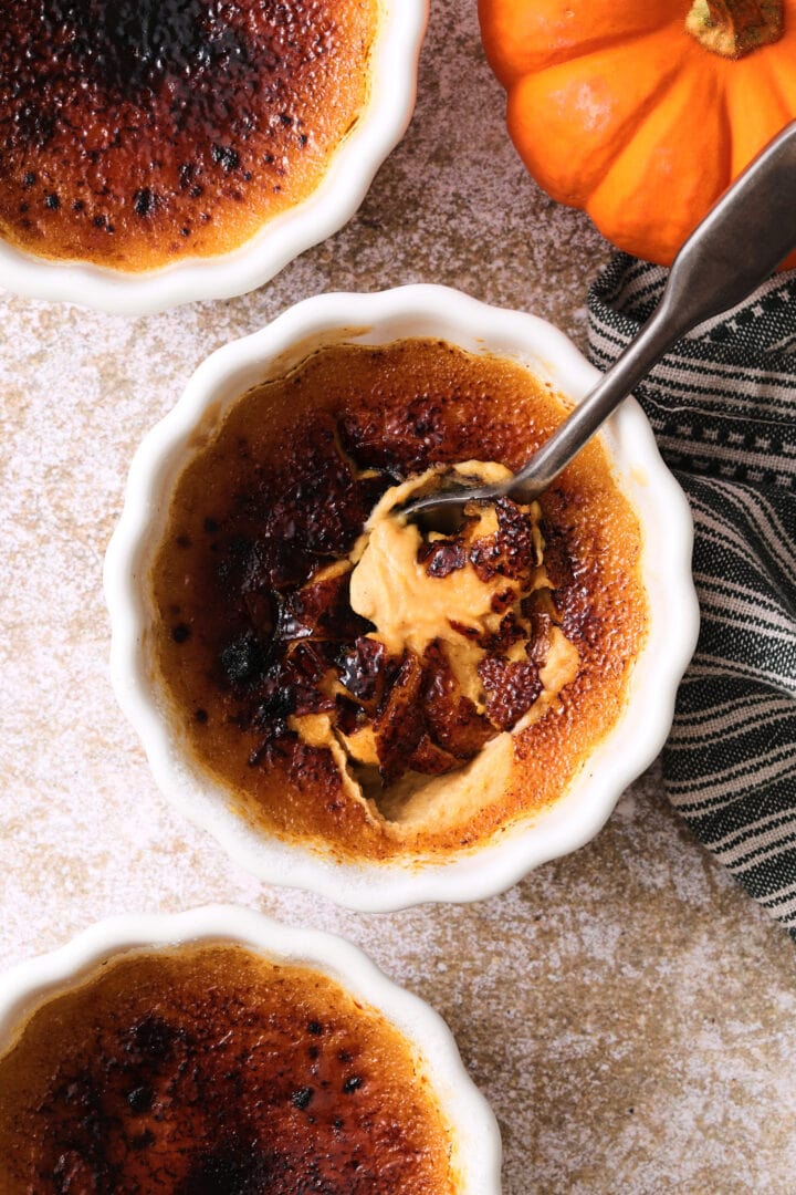 small white ramekin of pumpkin creme brulee, the burnt sugar crust has been cracked by a spoon and the pumpkin custard is just visible underneath.